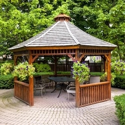 Brown wooden gazebo surrounded by hanging potted plants with bushes and trees on stone bricks